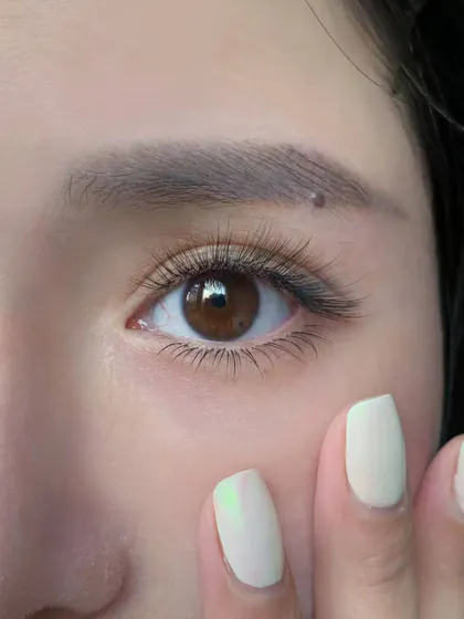 Close-up of an eye with a soft, natural-looking lash extension set, and fingertips with milky white gel nails resting on the cheek