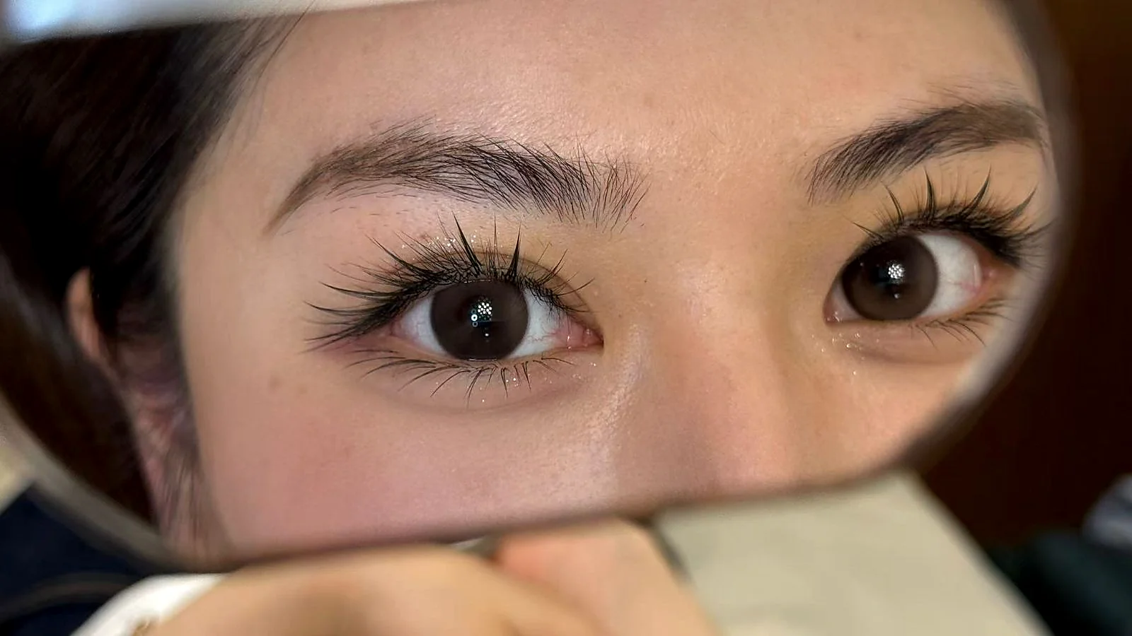 Through-mirror view of a client mid-session — one eye with the finished Zero-Sensation Ultra-Lock velvet lashes, the other eye still natural — same client, same chair